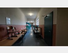 Ein langer, schlichter Schulflur mit beiger Wandfarbe im oberen Bereich und roter Farbe im unteren Bereich. Entlang der Wände stehen mehrere zusammengestellte Tische und Stühle mit hochgestellten Sitzflächen; rechts befindet sich eine dunkelgrüne Tür und links ein kleines Wandfach.