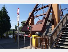Metallbrücke mit schrägen Verstrebungen und Treppenzugang, daneben eine Schranke mit rotem Lichtsignal und ein Schild „Radfahrer frei“. Im Hintergrund ein kleiner Wasserkanal, umgeben von Bäumen und Gebäuden.
