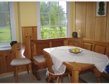 Ecke einer rustikalen Küche oder eines Esszimmers mit Holzmöbeln, darunter eine Eckbank und Stühle mit gepolsterten Sitzflächen. Auf dem Holztisch liegt eine Tischdecke, darauf steht eine Schale mit Äpfeln; im Hintergrund sind große Fenster mit Ausblick auf eine grüne Wiese.