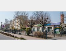 Straßenansicht mit einem eingeschossigen Gebäude, das vermutlich eine Kita oder Schule ist, erkennbar an farbigen Elementen und einer Regenbogen-Wandmalerei. Daneben befindet sich ein mehrstöckiges Wohnhaus, umgeben von kahlen Bäumen und einem umzäunten Hof.