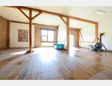 Ein geräumiger, heller Innenraum mit sichtbaren Holzbalken und Backsteinwänden, ausgestattet mit Holzboden. Der Raum enthält ein großes Fenster, einen blauen Sessel, eine Leiter und einen freistehenden Ofen.