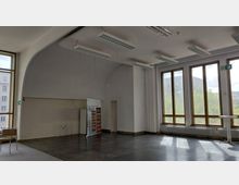 Ein leerer, heller Raum mit großen Fenstern, die viel Tageslicht einlassen. Der Raum hat dunkle Fliesenböden, eine abgerundete Decke, mehrere Hängelampen und einen Notausgang-Hinweis über einer Tür.