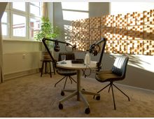Ein kleiner, moderner Podcast-Raum mit einem runden Tisch, zwei schwarzen Stühlen und Mikrofonarmen. Die Wand ist mit schallabsorbierenden Holzelementen dekoriert, und ein Fenster lässt Tageslicht herein; eine grüne Zimmerpflanze steht in der Ecke.