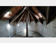 Dachboden eines alten Gebäudes mit sichtbarer Holzbalkenkonstruktion und einem mit Ziegeln gedeckten Dach. Die Wände sind aus grauem Putz, und durch kleine Öffnungen im Dach fällt Licht in den Raum.