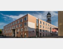 Modernes, mehrstöckiges Gebäude mit orangefarbener Fassade und rechteckigen Fenstern, im Hintergrund ragt ein Kirchturm mit Uhr und barocker Kuppel in den Himmel. Die Umgebung zeigt eine Straßenecke mit Schildern und einem geparkten Auto.