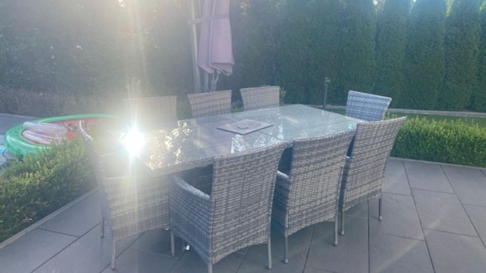 Terrasse mit großformatigen Steinplatten, einem rechteckigen Glastisch und sechs Korbstühlen; in der Mitte steht ein geschlossener Sonnenschirm. Im Hintergrund begrenzen eine niedrige Hecke und hohe dichte Nadelbäume den Gartenbereich.