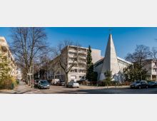 Straßenszene mit modernen Wohnhäusern links und einer Kirche mit spitz zulaufendem, weißem Turm und Kreuz rechts. Die Straße ist gesäumt von Bäumen ohne Laub und parkenden Autos vor einer klaren, blauen Himmelkulisse.