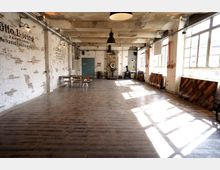 Industriehalle mit einer rustikalen Einrichtung, bestehend aus freiliegenden Backsteinwänden, Holzboden und großen Fenstern, die viel Tageslicht hereinlassen. Im Raum befinden sich einige Holztische und -stühle sowie historische industrielle Dekorationsobjekte wie alte Maschinen.