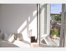 Helles Zimmer mit weißer Wand, einem Bett mit weißen Kissen und einem offenen Fenster, das auf eine Terrasse blickt. Auf der Terrasse befinden sich Pflanzen in Töpfen und ein Teil eines Gebäudes ist im Hintergrund sichtbar.