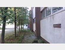 Außenbereich eines Gebäudes mit brauner und weißer Fassade, flankiert von Bäumen auf einem gepflasterten Weg. Im Hintergrund ist ein Hof mit Fahrradständern und einer weißen Tür sichtbar.