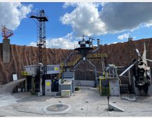 Industriell gestaltetes Außenareal mit Containern, einem Hubschrauberwrack und stählernen Konstruktionen, umgeben von einer hohen, rötlich-braunen Felswand. Im Vordergrund sind ein Betonplatz mit einem markierten Landeplatz und eine Antenneninstallation zu sehen.