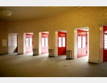 Heller Innenraum mit gelblichen Backsteinwänden und Betonboden, unterteilt durch mehrere rechteckige Durchgänge mit leuchtend roten Türen und Fensterelementen. Eine gewölbte Decke und schlichte Wandlampen rahmen den minimalistisch gestalteten Raum.