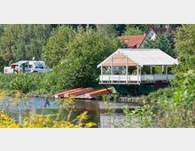 Ein Pavillon mit weißem Dach ragt direkt über ein Gewässer hinaus und dient als Sitzbereich, teils von Menschen besetzt. Im Hintergrund befinden sich ein Wohnwagen, ein Auto und dicht bewachsene Bäume; am Ufer liegen orangefarbene Boote.