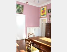 Esszimmer mit rosa gestrichenen Wänden, einem hölzernen Esstisch und Stühlen im Vordergrund. An den Wänden hängen verschiedene Kunstwerke, darunter ein buntes abstraktes Bild; ein weißer Heizkörper und eine hölzerne Kommode sind ebenfalls zu sehen.