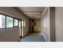 Kleiner Innenraum mit grobem, beigefarbenem Putz an den Wänden, einem großen Fenster- und Türbereich aus Holz auf der linken Seite und farbigen Zickzack-Streifen an der Stirnwand. Am Boden sind Spuren von Abnutzung sichtbar, und ein Feuerlöscher ist an der Wand montiert.