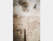 Decke und Wand eines verlassenen Gebäudes mit abgeblätterter Farbe, einem abgenutzten Geländer im Vordergrund und einer Deckenlampe mit geprägtem Glas.