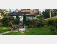 Ein Einfamilienhaus mit weißer Fassade und Walmdach, umgeben von einem gepflegten Garten mit Rasen, Sträuchern und hohen Zypressen. Im Vordergrund befindet sich eine Holzbank sowie ein kleiner gepflasterter Bereich mit Töpfen und einer dekorativen Vogeltränke.