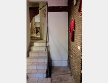 Ein schmaler Treppenaufgang mit weißen Fliesen führt zu einer Tür mit der Aufschrift „WC“. Rechts davon ist eine Ziegelwand mit einem aufgehängten roten Trikot zu sehen, daneben ein weißer Wandbereich und ein Holzfußboden im Vordergrund.