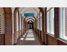 Langer Flur mit Backsteinbögen und großen Fenstern auf der rechten Seite, durch die Sonnenlicht auf den dunklen Boden fällt. Ein blaues Schild in der Mitte des Flurs weist den Weg zu "Gebäude C" und dem "Ausgang".