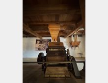 Historischer Innenraum mit einer alten landwirtschaftlichen Maschine aus Metall und Holz, wahrscheinlich eine Getreidemühle. Im Hintergrund sind eine Holzkonstruktion an der Decke, weiße Wände und ein schwarz-weißes Foto an der Wand zu sehen.