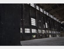 Innenansicht einer verlassenen Industriehalle mit hohen, dunklen Betonwänden und großen, rechteckigen Fenstern, die in Reihen angeordnet sind und wenig Licht hereinlassen. Die Halle wirkt leer und weist sichtbare Anzeichen von Verfall auf, wie beschädigte Wände und bröckelnden Putz.