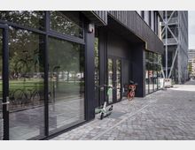 Moderne Gebäudefassade mit großen Glasfenstern und schwarzen Rahmen, die eine gepflasterte Straße und grüne Parkflächen im Hintergrund reflektieren. Im Vordergrund stehen ein E-Scooter und ein orangefarbenes Leihfahrrad, während ein angrenzendes Gebäude mit Stahlträgerkonstruktion sichtbar ist.