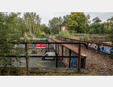 Ehemalige Eisenbahnbrücke mit verrostetem Geländer und überwucherten Schienen, darunter eine Straße mit einem weißen Lieferwagen und einem Fahrradfahrer vor einer Wand mit Plakaten und Graffiti, umgeben von dichter Vegetation.