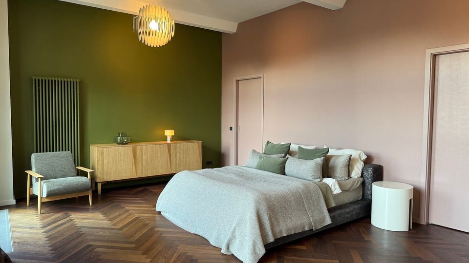 Modern bedroom with a low upholstered bed dressed in neutral linens, set on a dark herringbone wood floor. The room features a green accent wall with a radiator and wooden sideboard, a gray armchair, soft pink walls, and two sculptural pendant lights.