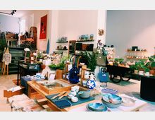 Ein moderner Laden mit einer Mischung aus handgefertigten Keramiken, Pflanzen, Taschen und anderen dekorativen Gegenständen. Die Einrichtung umfasst Holztische, Regale, Dekorationen aus Naturmaterialien und eine kleine Treppe im Hintergrund, die zu weiteren Warenbereichen führt.