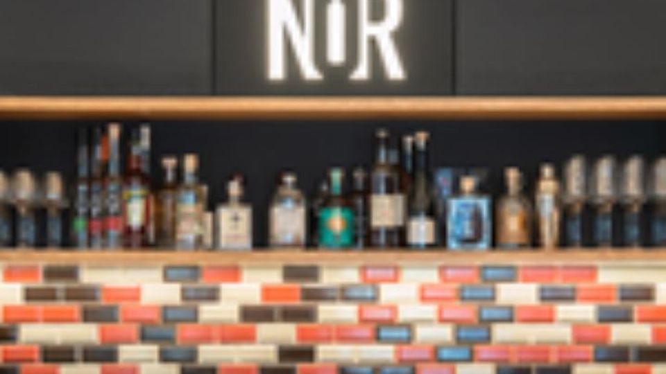 Innenansicht einer Bar mit einer langen Holzablage, auf der zahlreiche Spirituosenflaschen vor einer dunklen Wand stehen. Unter der Ablage befindet sich eine Wandverkleidung aus rechteckigen, mehrfarbigen Kachelsteinen in Rot-, Grau- und Beigetönen.