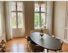 Esszimmer in einem Altbau mit großen Doppelfenstern, durch die Tageslicht einfällt, und Blick auf gegenüberliegende historische Gebäude. Ein ovaler Esstisch mit minimalistischer Dekoration steht auf einem Holzboden, umgeben von gepolsterten Stühlen.