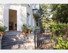 Eingangsbereich eines Wohnhauses mit offenstehender weißer Tür, die zu einem Innenraum führt. Vor der Tür befinden sich Pflanzentöpfe, eine Treppe aus Stein und ein schmiedeeisernes Gartentor, umgeben von Bäumen und Sträuchern.