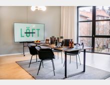 Moderner Konferenzraum mit einem langen Tisch, umgeben von schwarzen Stühlen, darauf Laptops, Getränkeflaschen, Gläser und Dekoration. Im Hintergrund ein großer Bildschirm mit der Aufschrift "LÖFT" sowie große Fenster mit Blick auf Wohnhäuser und Bäume.
