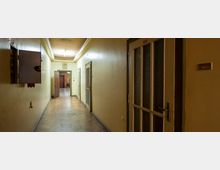 Ein schmaler, länglicher Flur mit cremefarbenen Wänden und Holzparkettboden. Rechts sind nummerierte Türen mit Glaseinsätzen, links ein offener Sicherungskasten; im Hintergrund ein geöffneter Durchgang und eine analoge Wanduhr über der Tür.