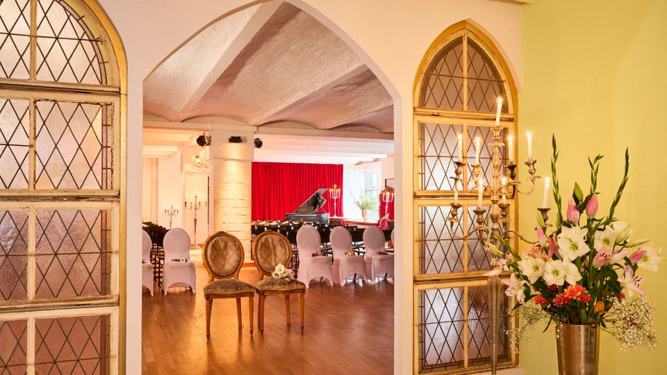 Blick durch einen Rundbogen in einen festlichen Veranstaltungssaal mit Holzboden, Stuhlreihen und einem Flügel vor roten Bühnenvorhängen. Links und rechts rahmen bleiverglaste Bogenfenster den Eingang; daneben stehen ein mehrarmiger Kerzenleuchter und eine große Blumenvase.