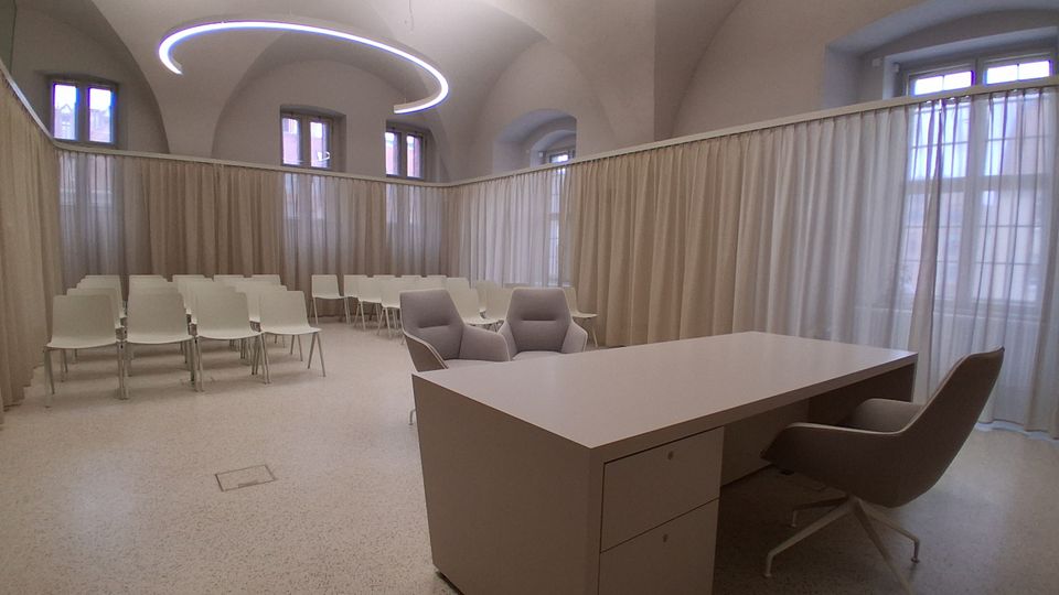 Heller Seminar- oder Besprechungsraum mit hohen Gewölbedecken, großen Fenstern und umlaufenden, halbtransparenten Vorhängen als Raumteiler. Im Vordergrund steht ein Schreibtisch mit Polstersesseln, dahinter mehrere Reihen weißer Stühle; an der Decke hängt eine ringförmige Leuchte.
