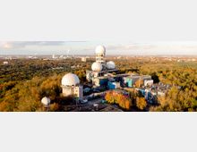 Luftaufnahme einer ehemaligen Radar- und Abhörstation im Wald, mit mehreren großen weißen Radarkuppeln (Radomen) auf flachen, teils verfallenen Gebäuden. Die Anlagen sind mit Graffiti bedeckt und von herbstlich gefärbten Bäumen umgeben; am Horizont ist eine weitläufige Stadtlandschaft zu sehen.