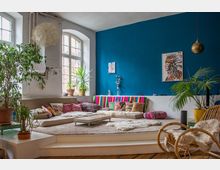Wohnzimmer mit erhöhtem Sitzbereich, blauer Akzentwand und großen Fenstern. Der Raum ist mit bunten Kissen, Teppichen, Pflanzen in Töpfen und einem hängenden goldenen Lampenschirm dekoriert.