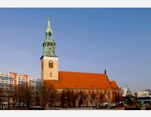 Außenansicht einer historischen Kirche mit rotem Ziegeldach und hohem, grün patiniertem Kirchturm, umgeben von Bäumen und modernen Gebäuden im Hintergrund unter klarem blauen Himmel.