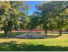 Ein Park mit einer Grünfläche, mehreren Bäumen und einer gepflasterten Fläche mit Sitzbänken im Vordergrund. Im Hintergrund verläuft eine rote Backsteinmauer, die den Bereich begrenzt.
