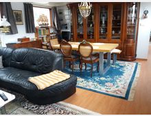 Wohnbereich mit einer schwarzen Ledercouch, einem Esstisch aus Holz mit Stühlen und einem blauen Teppich mit floralem Muster. Im Hintergrund stehen eine große, hölzerne Vitrine mit Glastüren und Regale mit Büchern sowie Dekorationsgegenständen.