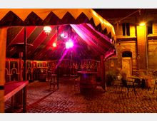 Zelt mit mittelalterlichem Dekor und rot-gelbem Streifenmuster auf einem gepflasterten Hof, beleuchtet von warmem gelbem Licht. Im Hintergrund ist eine Backsteinmauer mit Fenstern sowie Tische und Stühle im Außenbereich zu sehen.