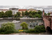 Flusslandschaft mit einer Steinbrücke auf der rechten Seite und einem Wohngebiet im Hintergrund. Im Vordergrund fließt das Wasser vorbei an Vegetation und einem kleinen Boot, während ein Anlegesteg und mit Graffiti bedeckte Ufermauern sichtbar sind.
