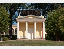 Kleiner klassizistischer Pavillon mit vier weißen Säulen und einem gelben Außenbau, gekrönt von einem blau-weiß gestreiften Dachvorsprung. Eingebettet in eine parkähnliche Umgebung mit Bäumen und Rasen.