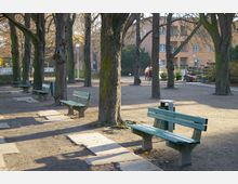 Ein kleiner Park mit einer Reihe von Bäumen, grünen Holz-Sitzbänken und einem sandigen Boden. Im Hintergrund sind ein Wohngebäude und ein Blumenstand zu sehen, neben Spaziergängern auf einem Weg.