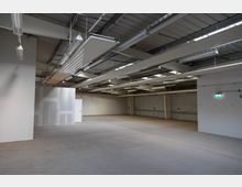 Großer, leerer Lagerraum mit glattem Betonboden, einer abgehängten Decke mit langen, parallel verlaufenden Leuchtstofflampen und sichtbarer Metallkonstruktion. An den Wänden befinden sich vereinzelt Wandanschlüsse und eine grüne Notausgangsbeschilderung.