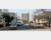 Straßenszene in einem Wohngebiet mit mehrstöckigen Wohngebäuden, einem hohen, gelben Wohnblock im Hintergrund und niedrigeren Gebäuden an den Seiten. Im Vordergrund sind parkende Autos, ein weißer Transporter, ein Straßenschild und ein Baum in einer bepflanzten Grünfläche zu sehen.