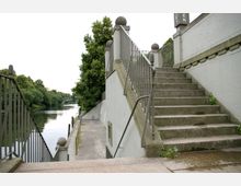 Steintreppe mit Geländern an beiden Seiten, die zu einem erhöhten Gehweg führt, parallel zu einem Fluss mit ruhigem Wasser und grünen Bäumen am Ufer; im Hintergrund ist eine kleine Brücke zu sehen.