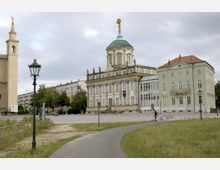 Historischer Platz mit barockem Gebäude, gekrönt von einer vergoldeten Atlas-Statue, flankiert von einem weiteren historischen Gebäude und einer Kirche mit Turm. Im Vordergrund führt ein Weg durch eine Rasenfläche, umgeben von Laternen.