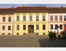 Gelbes, zweistöckiges historisches Gebäude mit roten Fensterrahmen und dekorativen Stuckelementen, flankiert von ähnlichen Gebäuden in Pastellfarben; im Vordergrund ein gepflasterter Platz mit einzelnen Pflanzkübeln.